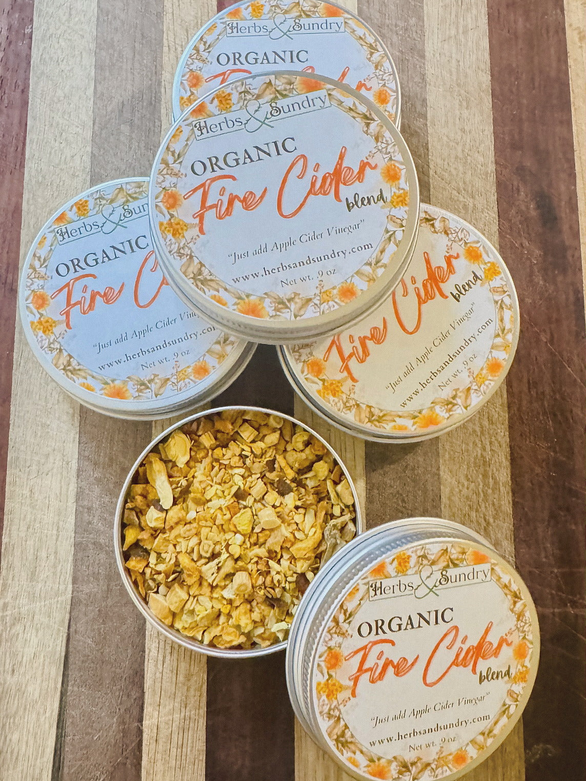 Stacked Organic Fire Cider Blend tins with orange labels, one open to show the golden dried roots and peel inside