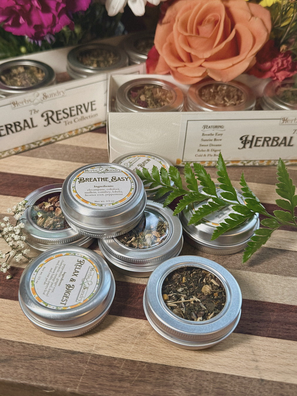 Herbal Reserve tins with fern fronds and flowers arranged on a striped wooden board