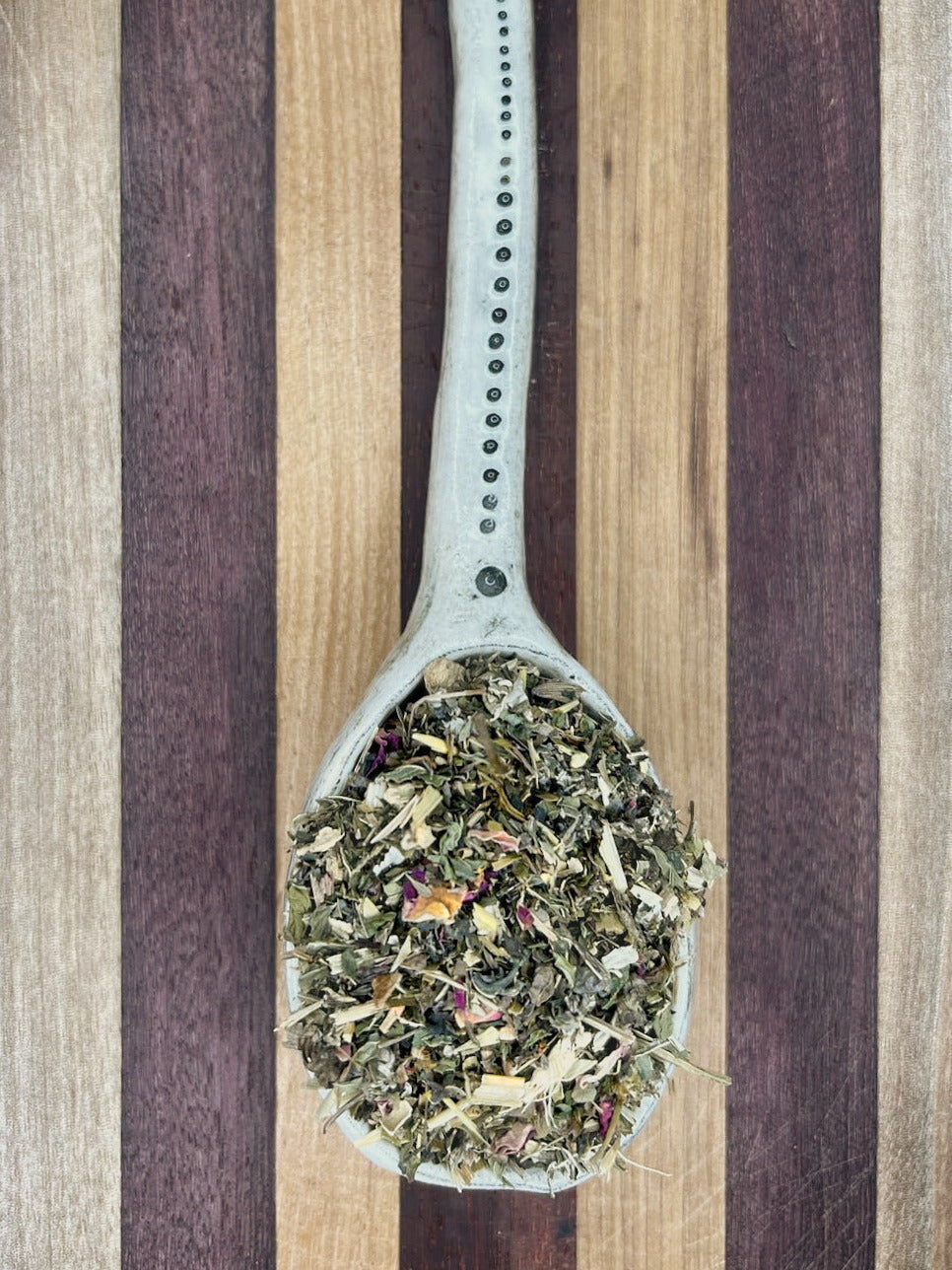 Pale blue spoon holding the green Mama To Be herbal blend on a striped wooden board