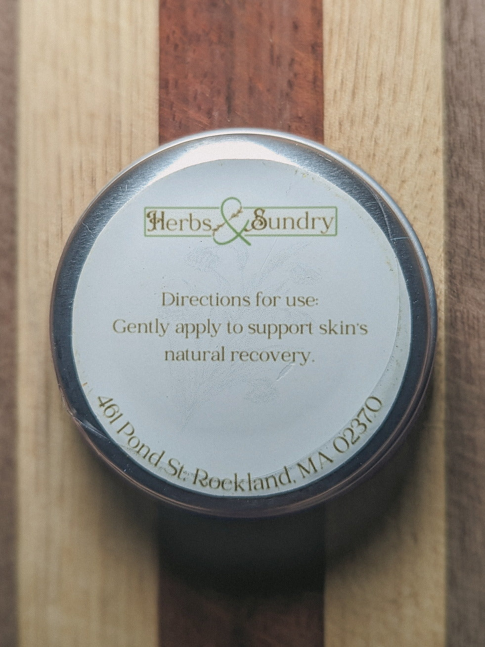 Underside of the Healing Comfrey Salve tin showing the directions label on a striped wooden board