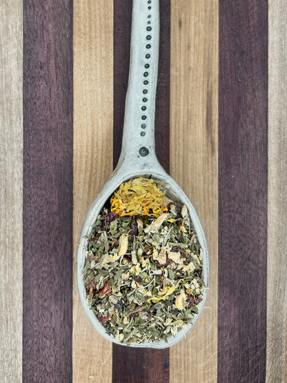 Pale blue spoon holding the Cold and Flu Brew blend, green herbs flecked with yellow elder flower and calendula