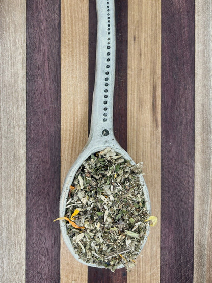Pale blue spoon holding the pale green Breathe Easy mullein blend on a striped wooden board