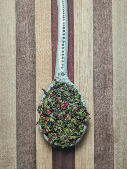 Pale blue spoon holding the Cool-Aid blend, deep red hibiscus petals mixed with green lemon balm and nettle